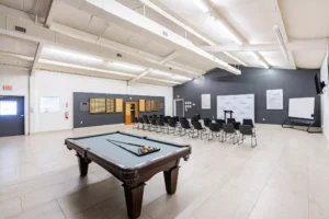 Spacious multipurpose room with rows of chairs set up for a group session, a pool table in the foreground, a whiteboard, and motivational posters on the walls.