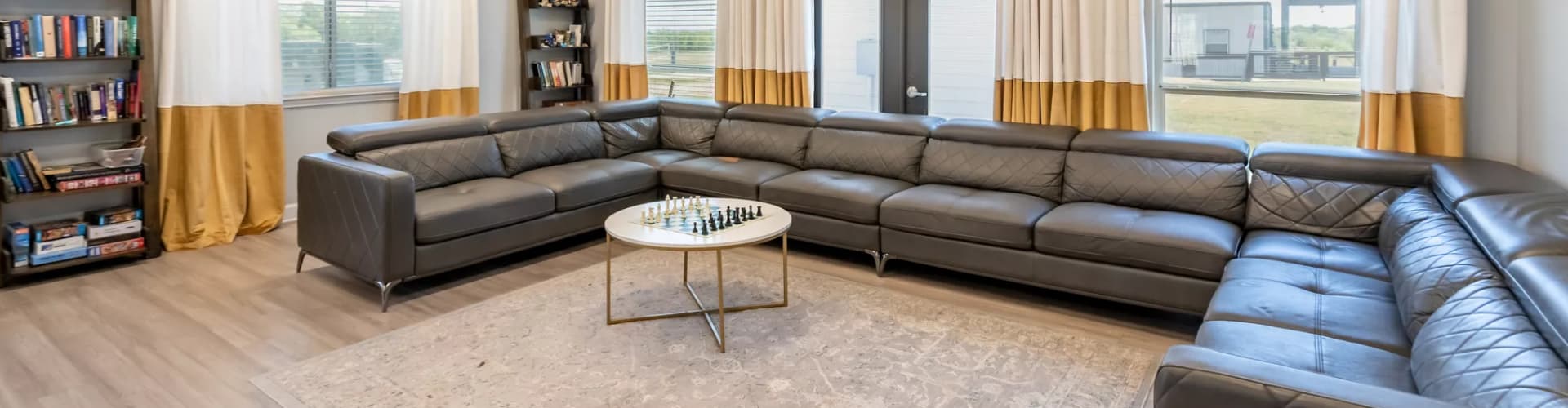 Spacious lounge area with large gray sectional sofas, a round coffee table with a chess set, bookshelves, and windows with white and gold curtains.
