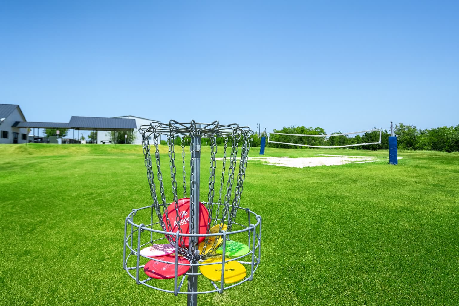 Outdoor recreation area with a disc golf basket filled with colorful discs and a sand volleyball court on a grassy field under a clear blue sky.