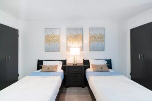 Clean, modern bedroom with two twin beds separated by a dark nightstand with a lamp, decorative pillows, and three gold and silver abstract paintings on the wall.