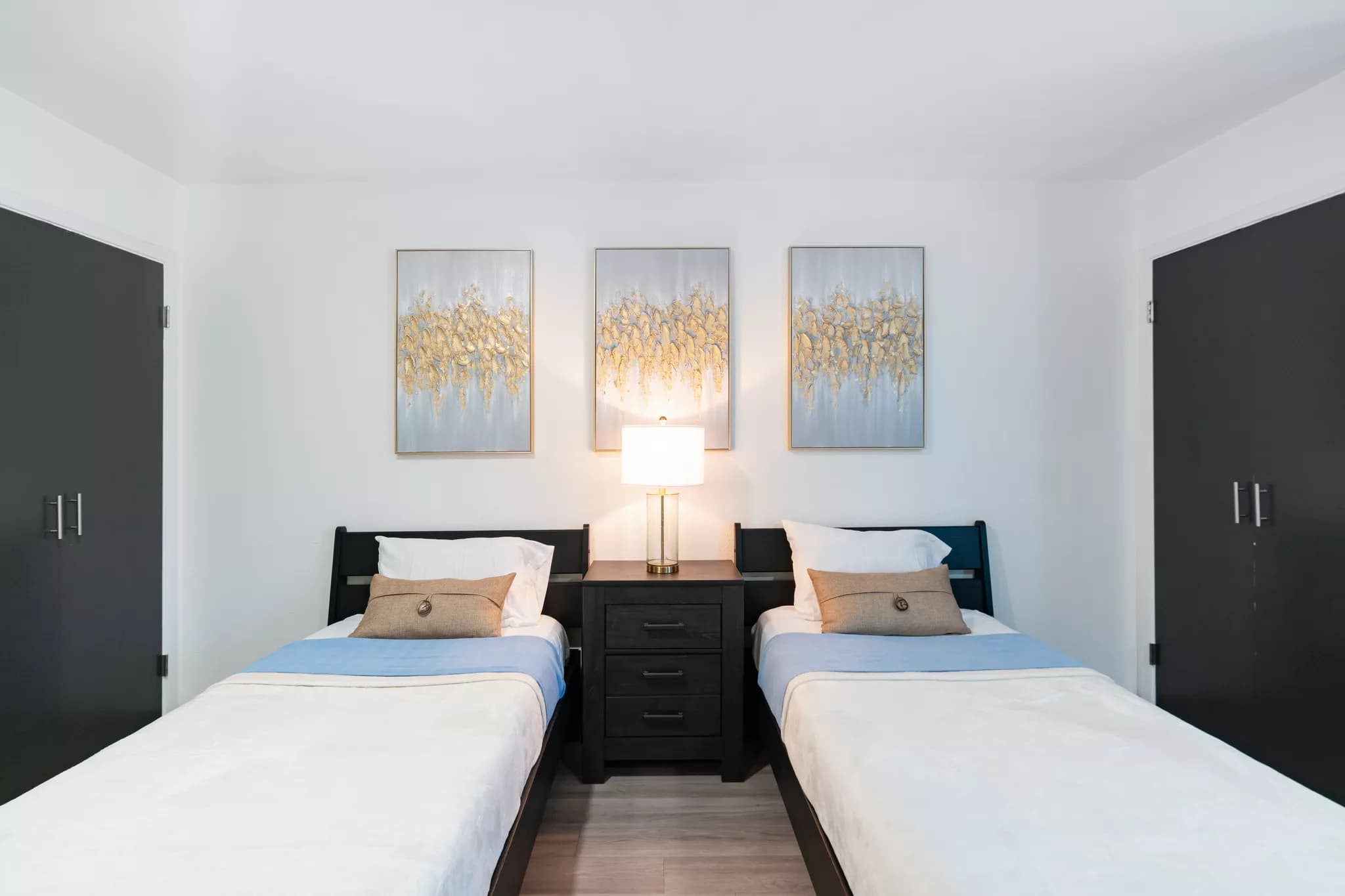 Clean, modern bedroom with two twin beds separated by a dark nightstand with a lamp, decorative pillows, and three gold and silver abstract paintings on the wall.