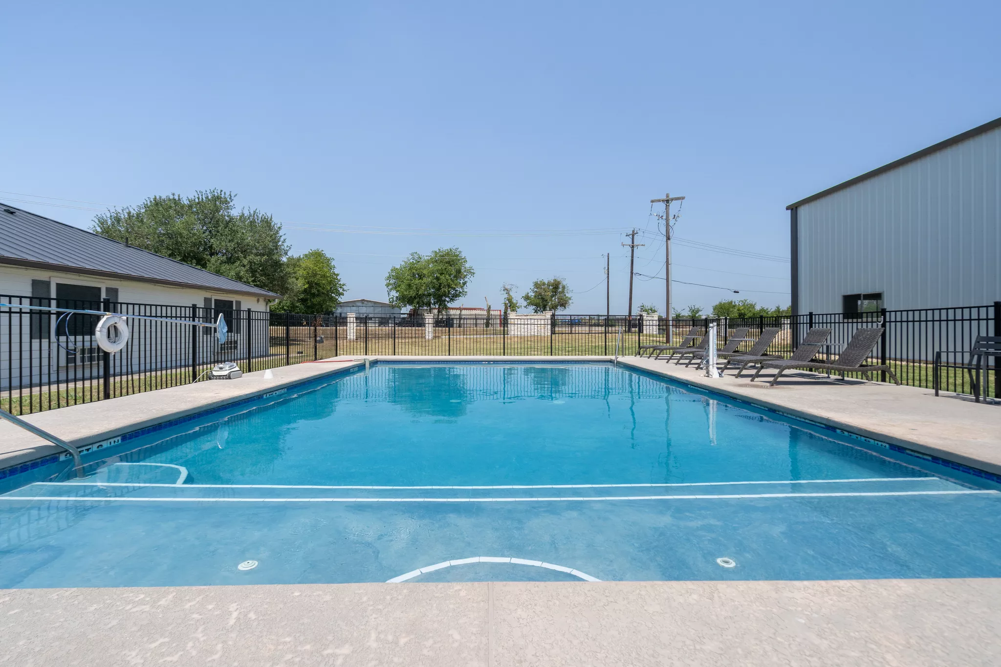 Outdoor swimming pool surrounded by a black safety fence with lounge chairs on the deck under a clear blue sky.