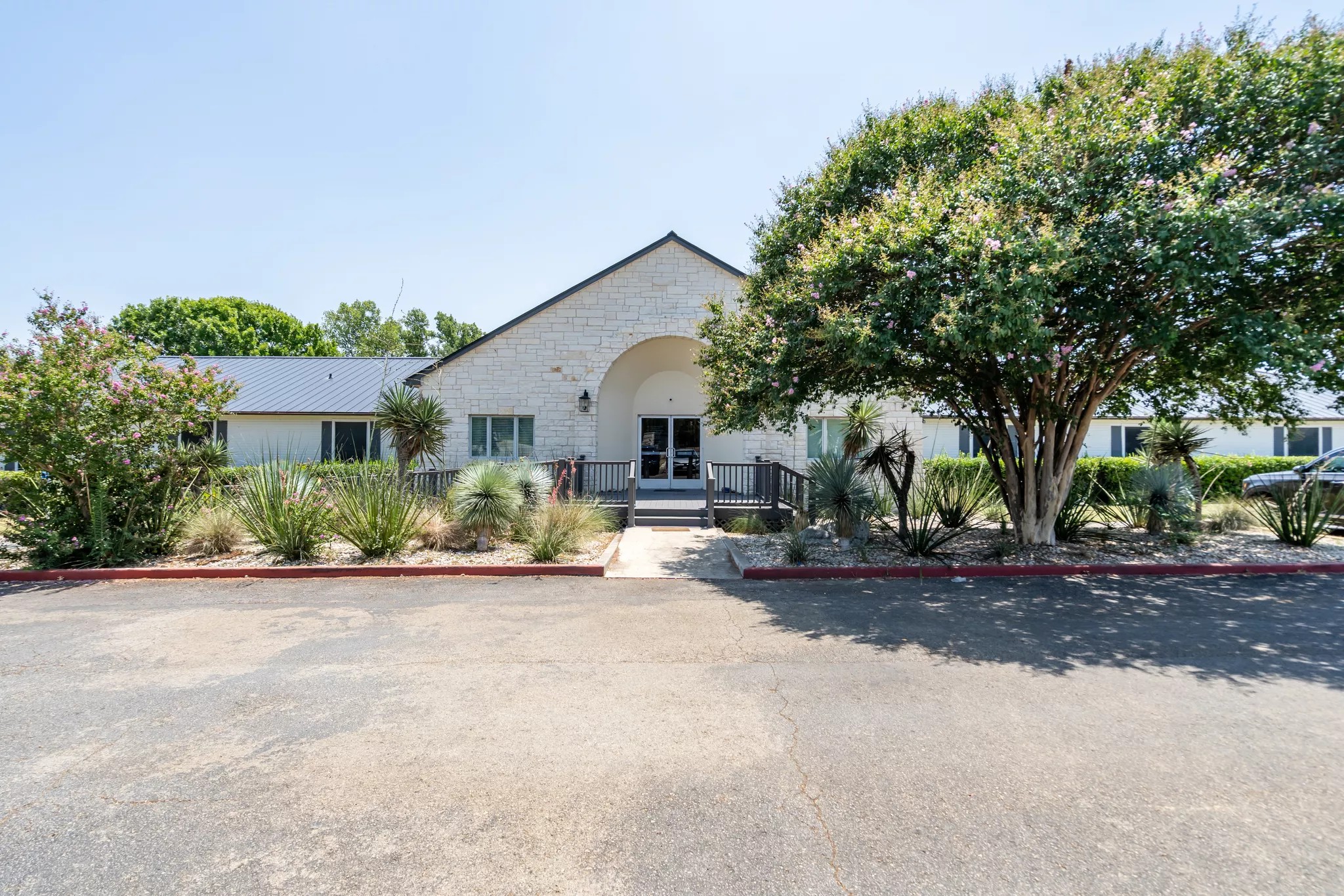 Front view of a light stone building with a peaked roof, double glass doors, ramp entrance, and surrounding desert landscaping with trees and plants.