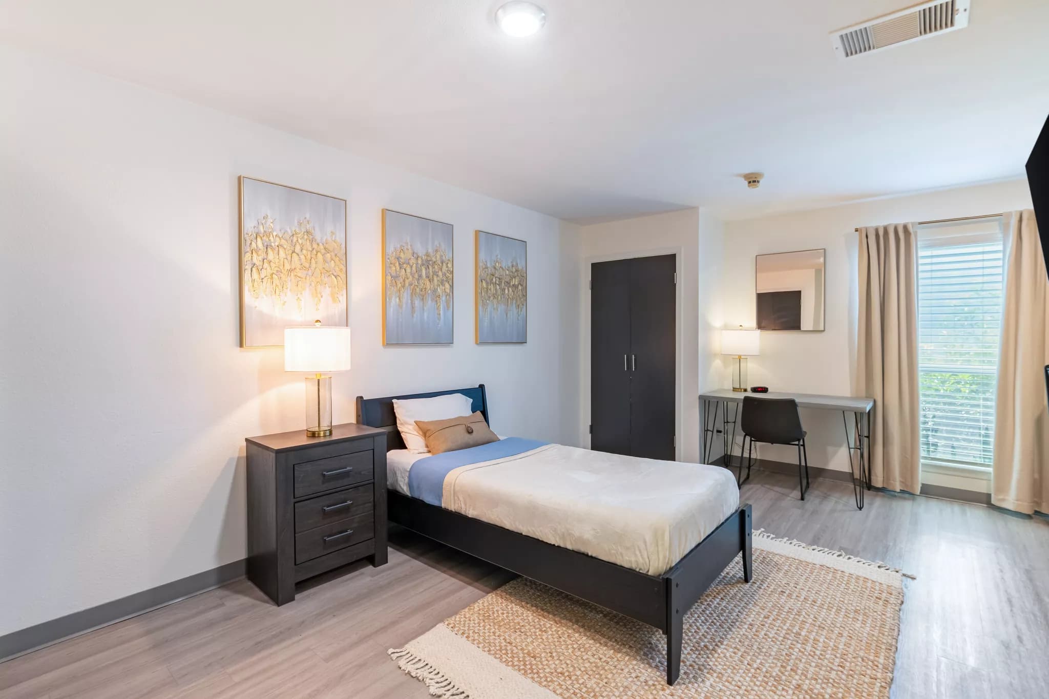 Clean, modern bedroom with one twin bed and a dark nightstand with a lamp, decorative pillows, and three gold and silver abstract painting on the wall