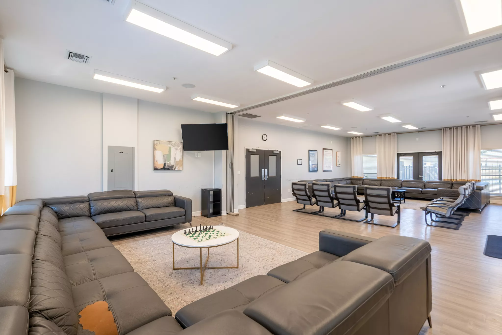 Spacious common room with black leather sofas and chairs arranged for group seating, a wall-mounted TV, framed artwork, and a round table with a chess set on a rug, under bright ceiling lights and large windows with curtains.