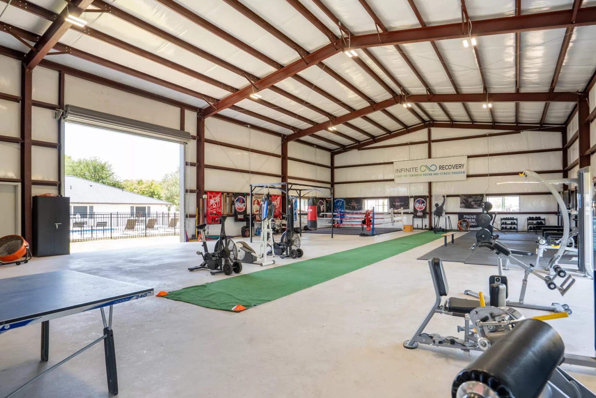 Spacious indoor gym with a boxing ring, punching bags, cardio and strength training equipment, green turf strip, and a banner reading ‘Infinite Recovery: Fight for Tomorrow Boxing and Fitness Program,’ with large open doors letting in natural light.