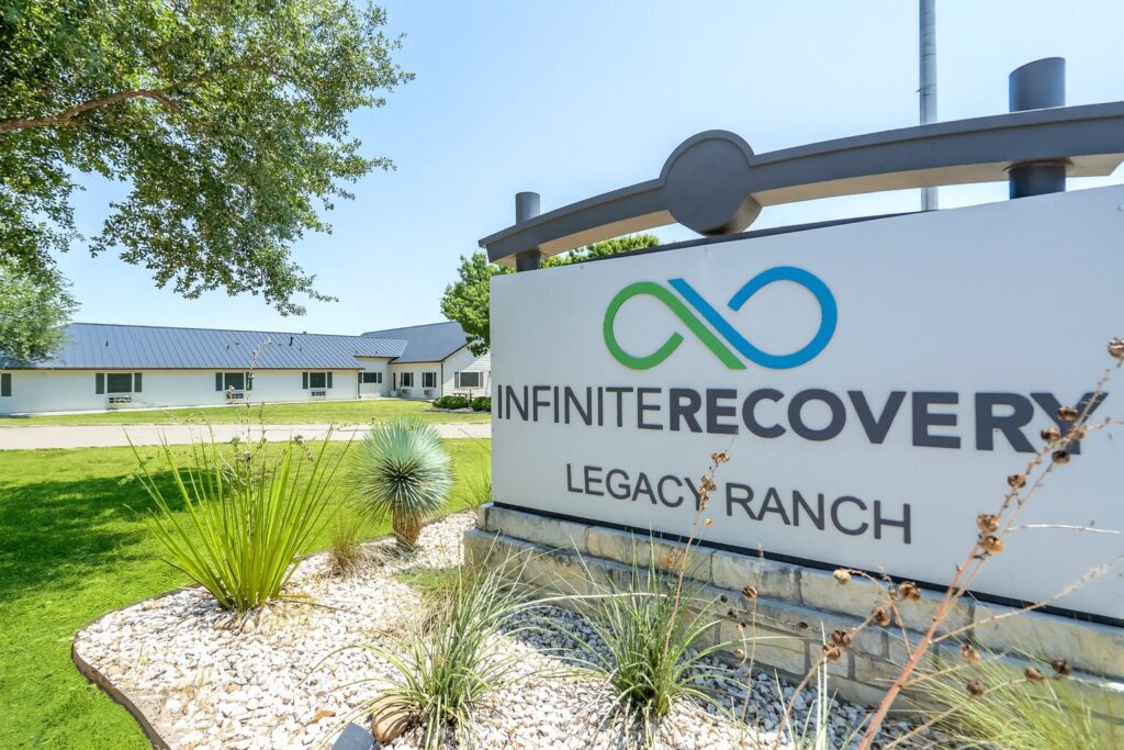 Exterior view of Infinite Recovery Legacy Ranch sign with logo in front of a white building and green landscaped lawn.