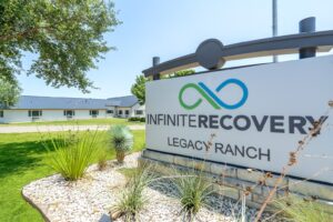 Exterior view of Infinite Recovery Legacy Ranch sign with logo in front of a white building and green landscaped lawn.