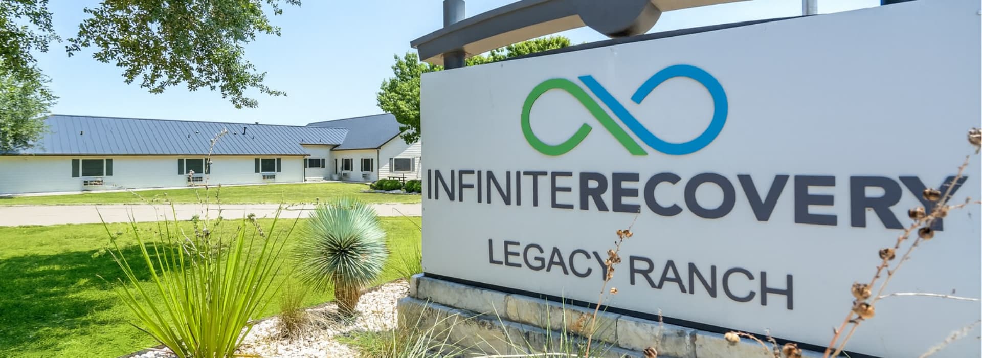 Exterior view of Infinite Recovery Legacy Ranch sign with logo in front of a white building and green landscaped lawn.