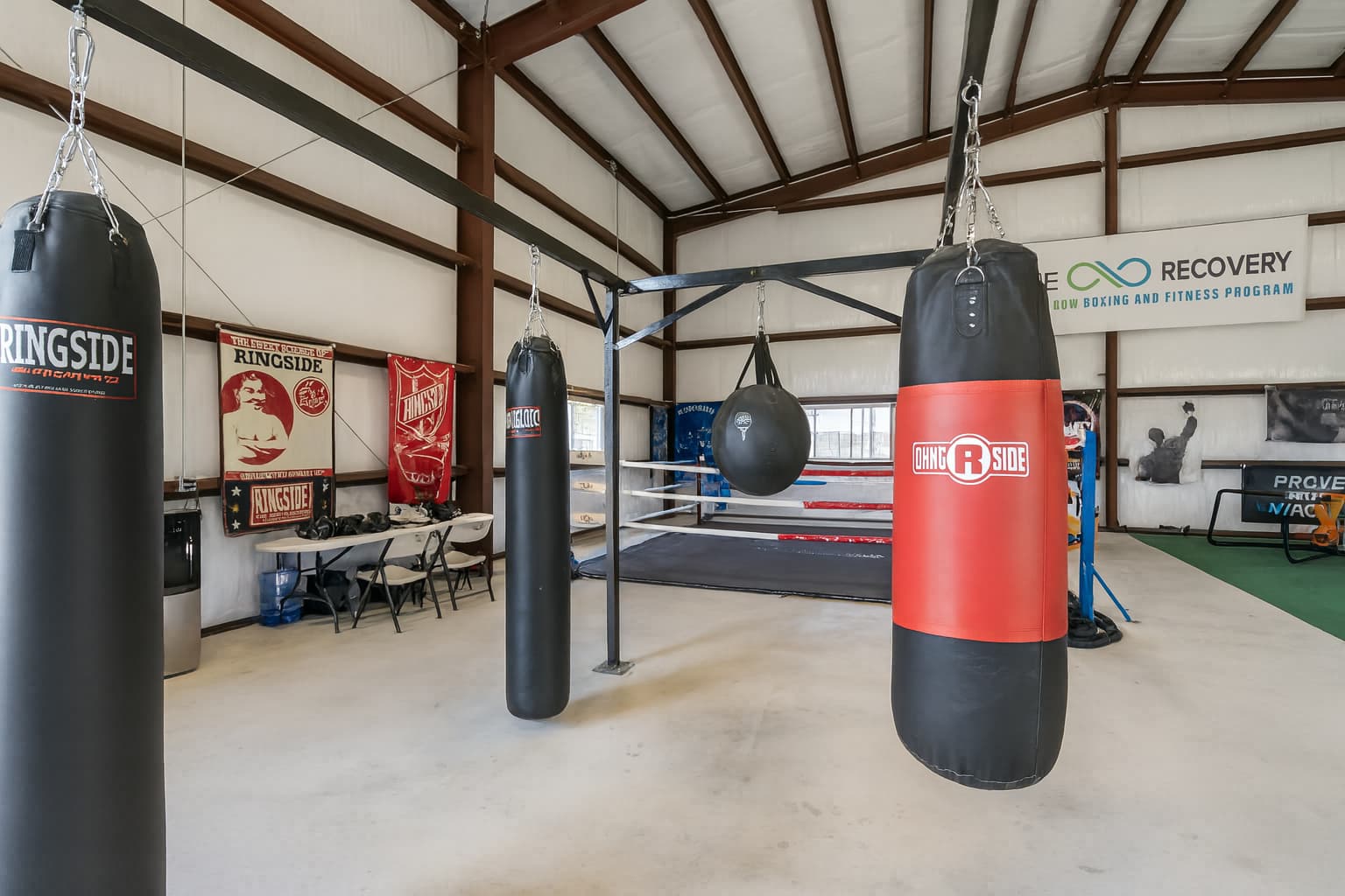 Indoor boxing gym with hanging punching bags, a boxing ring, workout mats, and training equipment inside a large metal building with exposed beams and fitness program banners on the walls.
