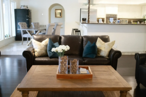 Cozy living room with a dark leather sofa decorated with gold and teal pillows, a wooden coffee table with vases and flowers, and an open view into the dining area and kitchen.