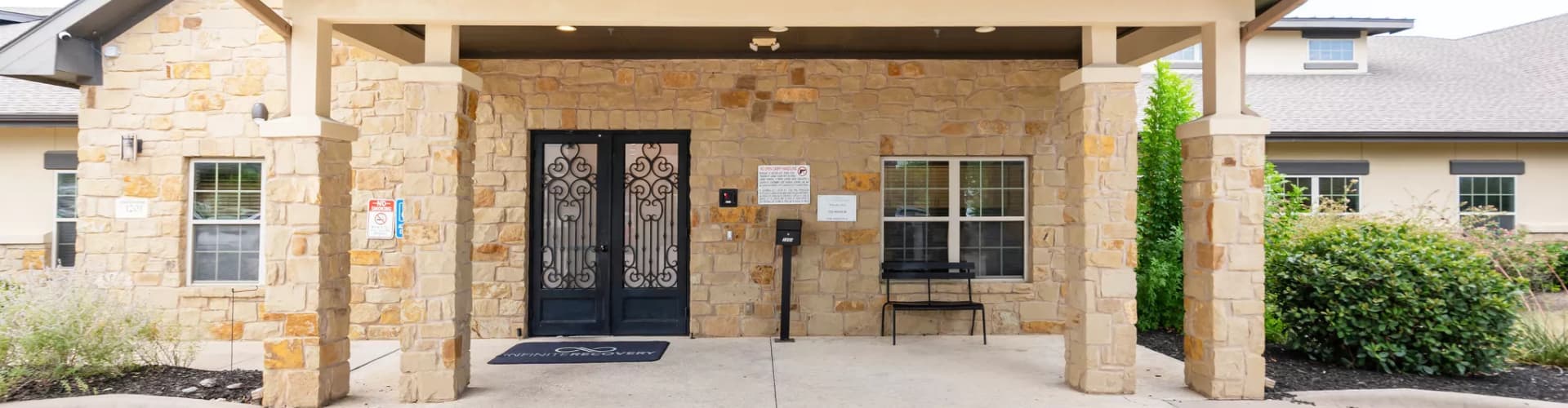 Front entrance of a stone building with decorative black double doors, covered porch, and surrounding greenery.
