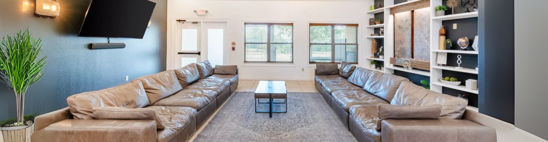Modern lounge with two long brown leather sofas facing each other, a central coffee table on a patterned rug, built-in shelving with decor, and a wall-mounted TV.