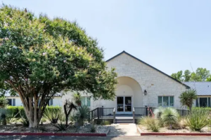 Front view of a light stone building with a peaked roof, double glass doors, ramp entrance, and surrounding desert landscaping with trees and plants.