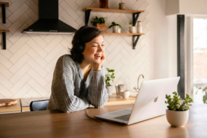 Smiling woman wearing headphones sits at a kitchen counter looking at her laptop, with potted plants and shelves in the background.