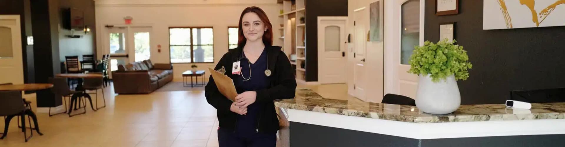 Smiling healthcare professional holding a clipboard and wearing scrubs stands at a reception desk in a bright, welcoming lobby with seating and decor.