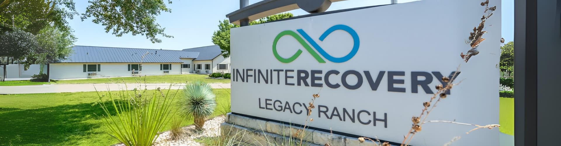 Exterior view of Infinite Recovery Legacy Ranch sign with logo in front of a white building and green landscaped lawn.