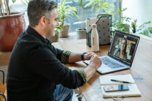 Man sitting at a wooden desk smiling during a video call on his laptop, with a notebook, phone, and water bottle nearby and plants in the background.