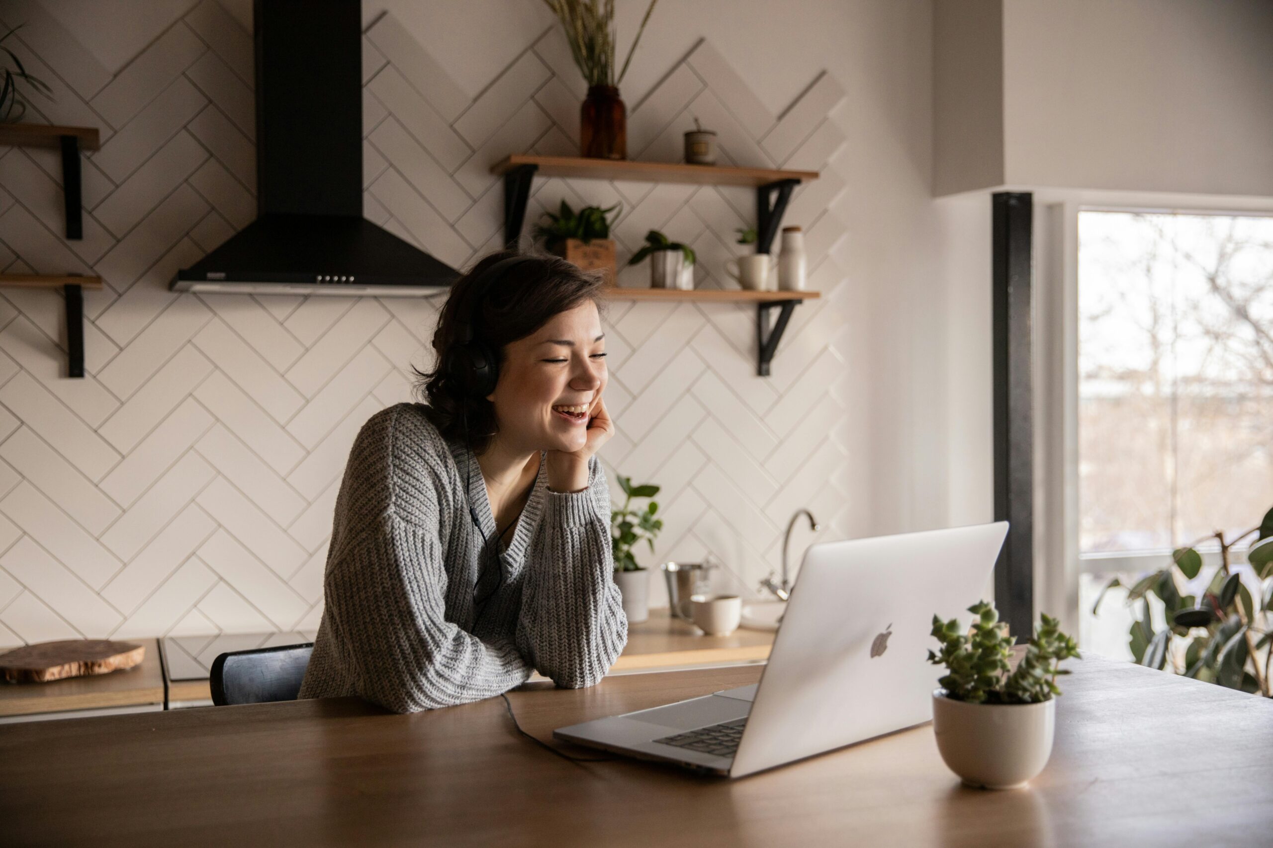 Smiling woman wearing headphones sits at a kitchen counter looking at her laptop, with potted plants and shelves in the background.