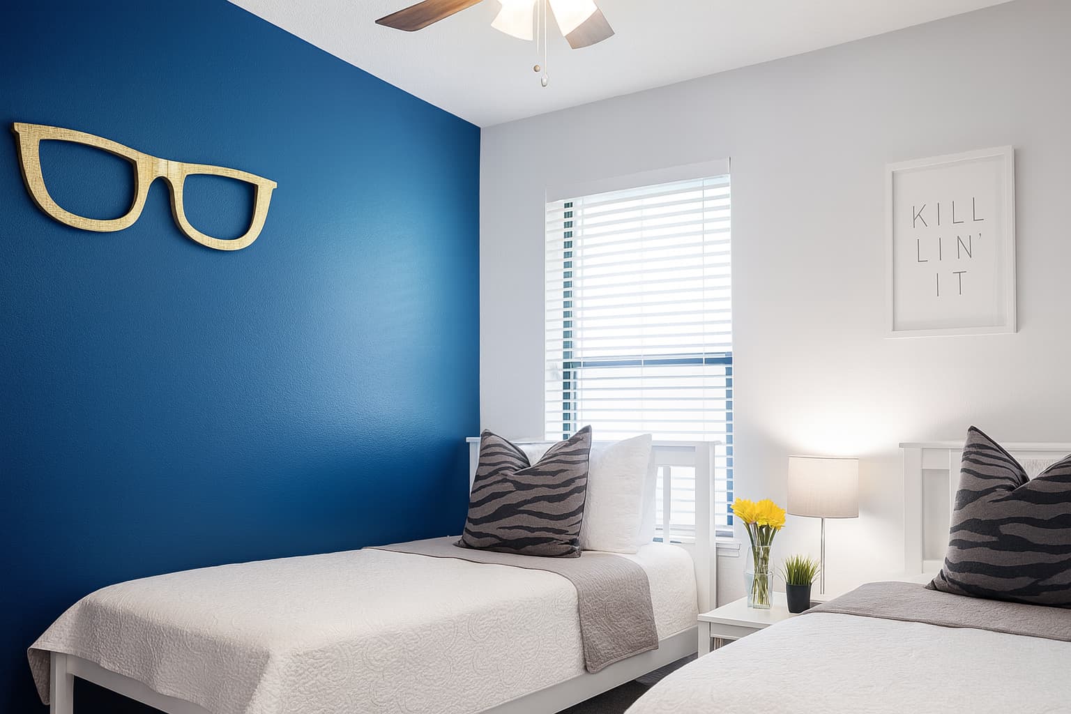 Bright bedroom with two twin beds, white bedding, zebra-print pillows, a blue accent wall with glasses decor, a nightstand with yellow flowers, and a framed sign reading ‘Killin’ It.’