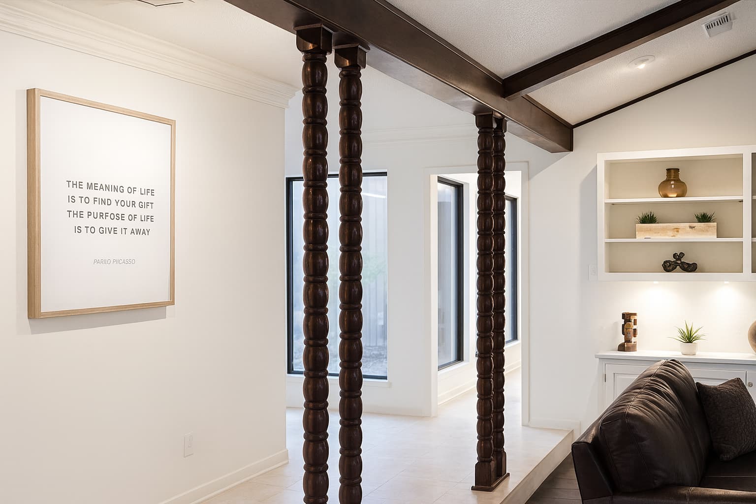 Modern living space with dark wooden beams and columns, a framed quote by Pablo Picasso on the wall, built-in shelving with decorative items, and a portion of a dark sofa.