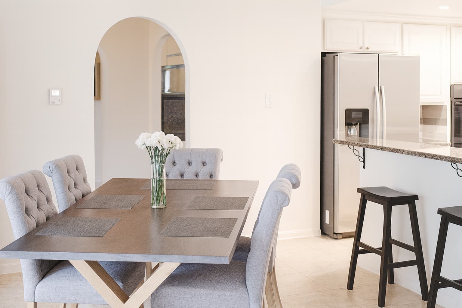 Bright kitchen and dining area with a wooden table, gray upholstered chairs, a vase of white flowers, and a granite breakfast bar with two black stools beside a stainless steel refrigerator.