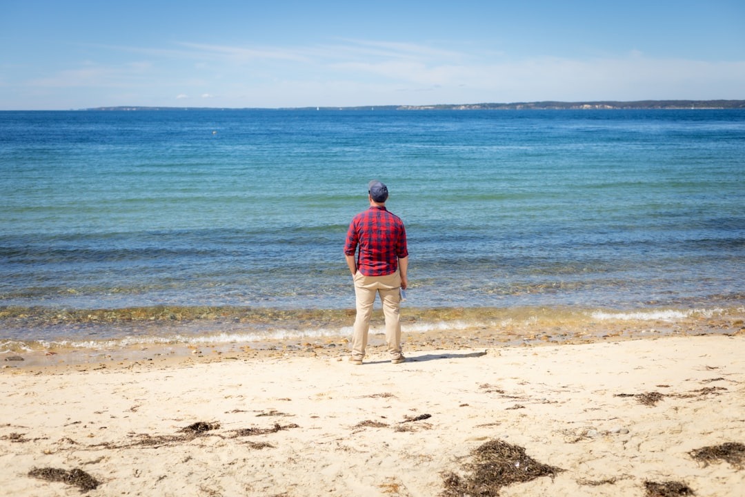 A man in a red flannel shirt stands on a sandy beach, looking out at the calm blue ocean during his recovery journey.