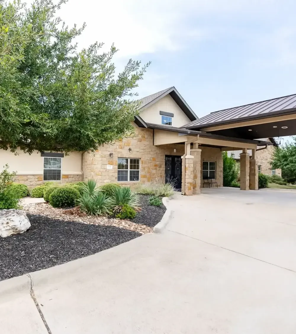 Exterior of a modern stone-and-stucco building with a covered entry supported by columns, a concrete driveway, and landscaped shrubs and mulch beds.