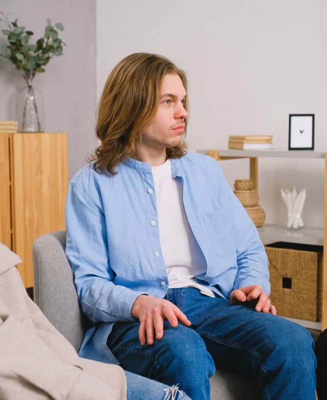 A man with shoulder-length brown hair sits on a gray couch, wearing a light blue button-up shirt and jeans, looking to the side.